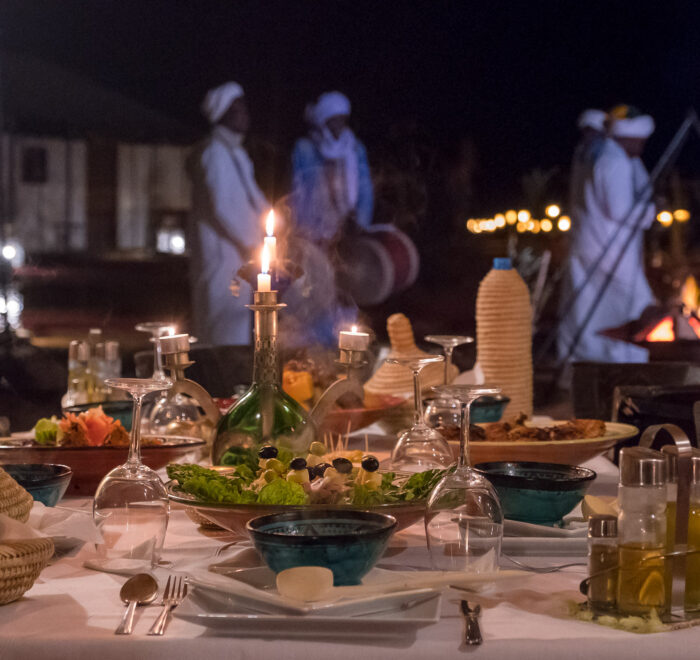 Gnawa musicians performing traditional music in the Sahara Desert, playing drums beside a table filled with Moroccan food.