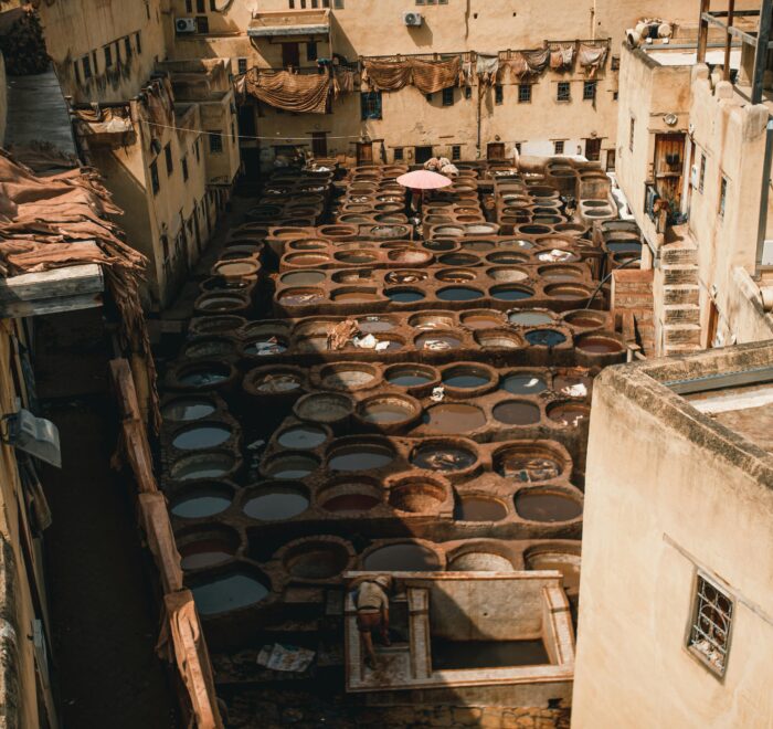 Overhead view of the Chouara Tanneries in Fes, Morocco, showing colorful dyeing vats surrounded by traditional stone buildings where leather is processed by skilled artisans.