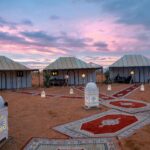 A cluster of traditional desert tents set up in the vast golden sands of the Moroccan Sahara under a clear blue sky, capturing the serene and remote beauty of the desert landscape.