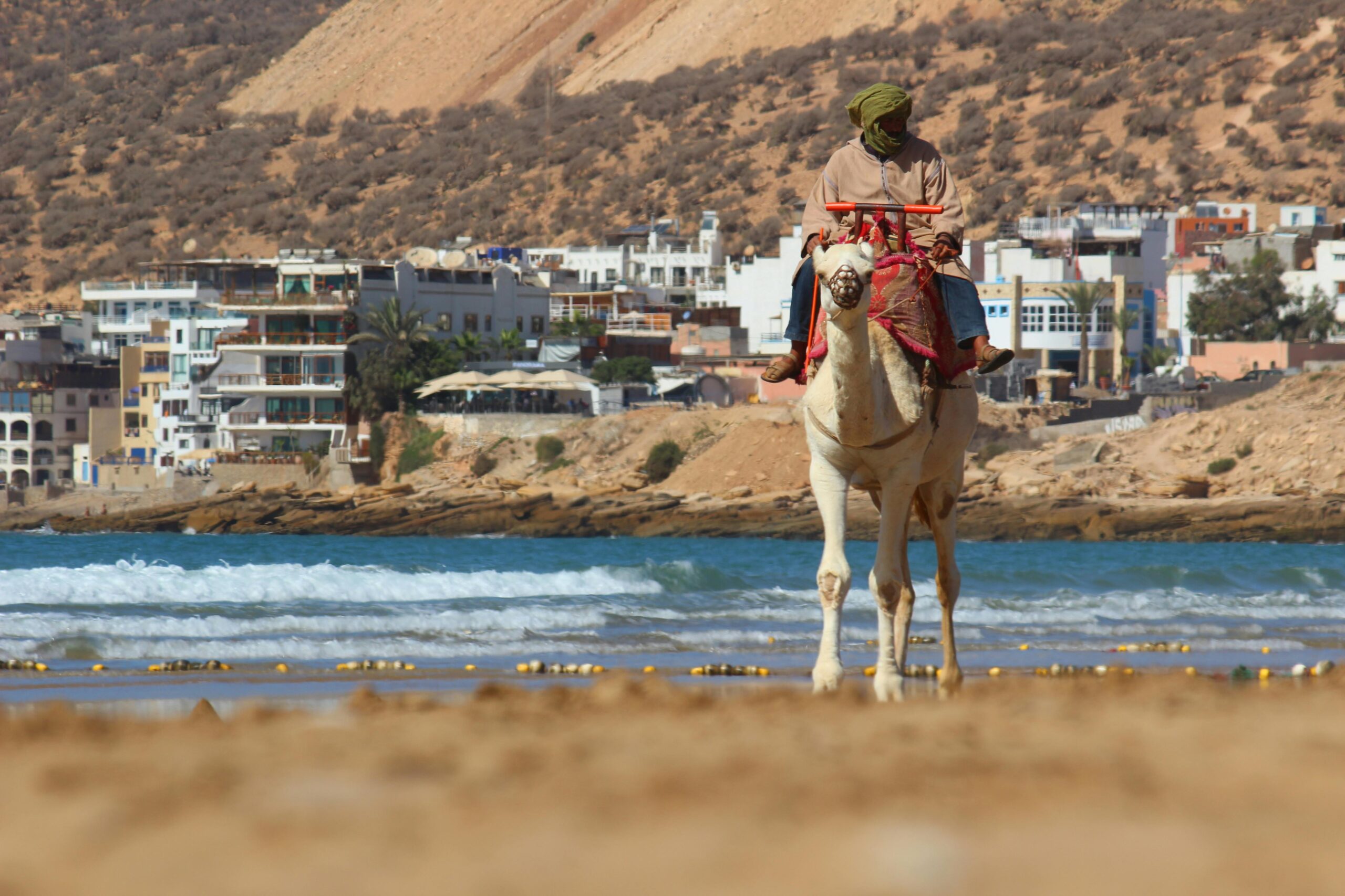 Man riding a camel along Taghazout Beach near Agadir, Morocco, with golden sands and Atlantic Ocean waves in the background.
